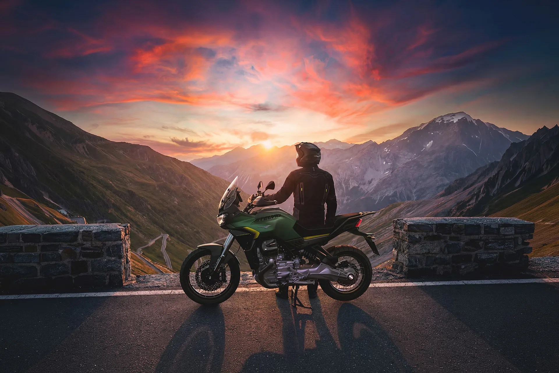 Person in black riding gear standing beside a green adventure motorcycle on a mountain road at sunset.