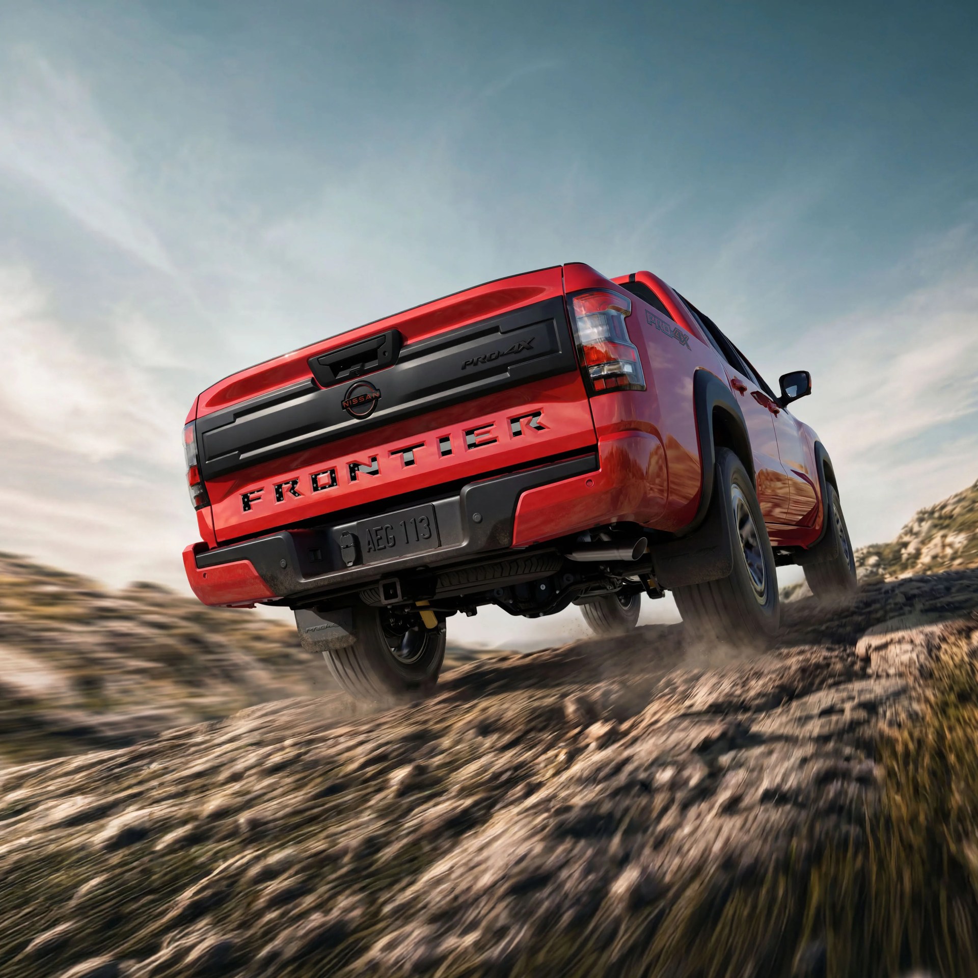 Red Nissan Frontier Pro-4X pickup truck driving off-road on rocky terrain under a blue sky.