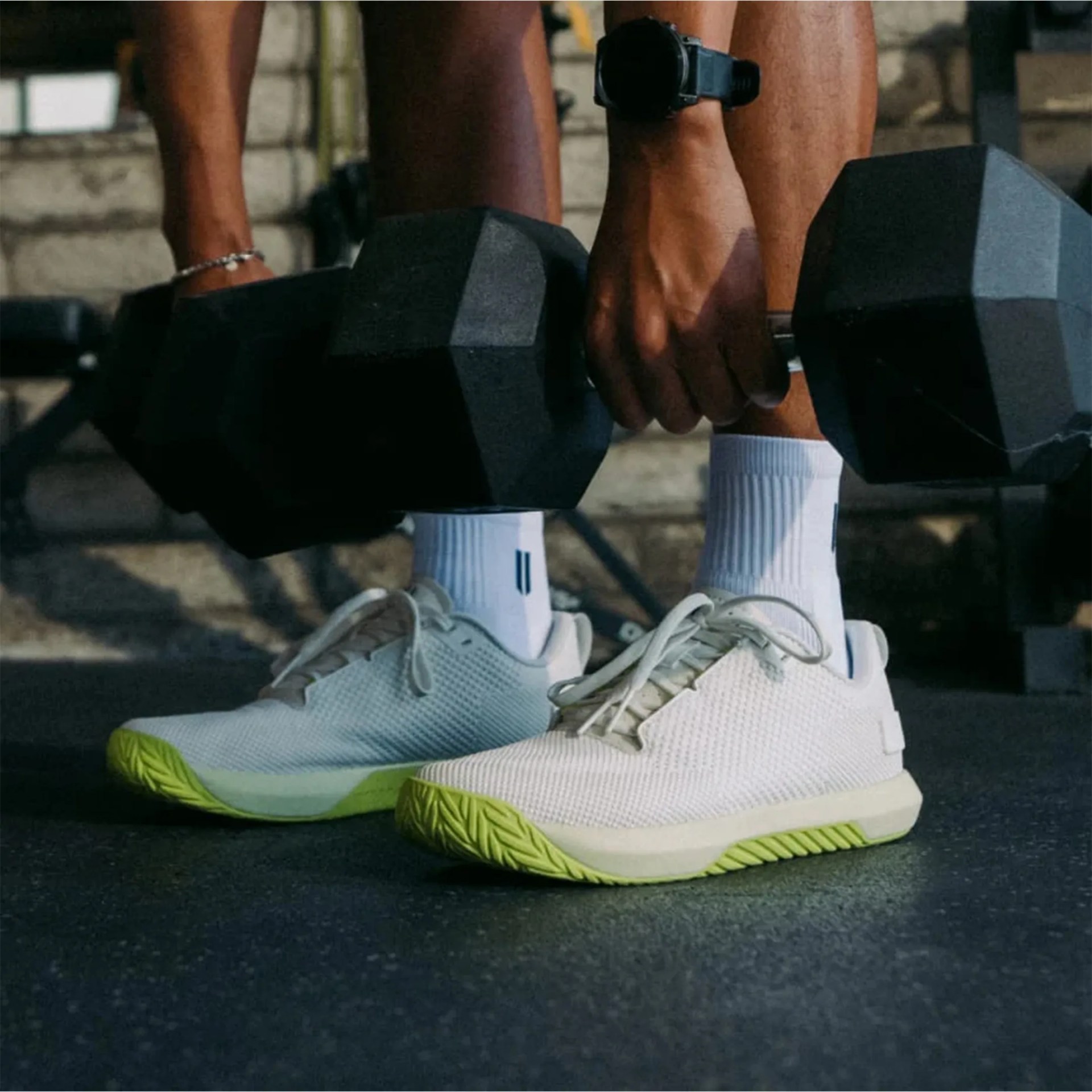 White athletic shoes with lime green soles worn by a person holding black hex dumbbells.