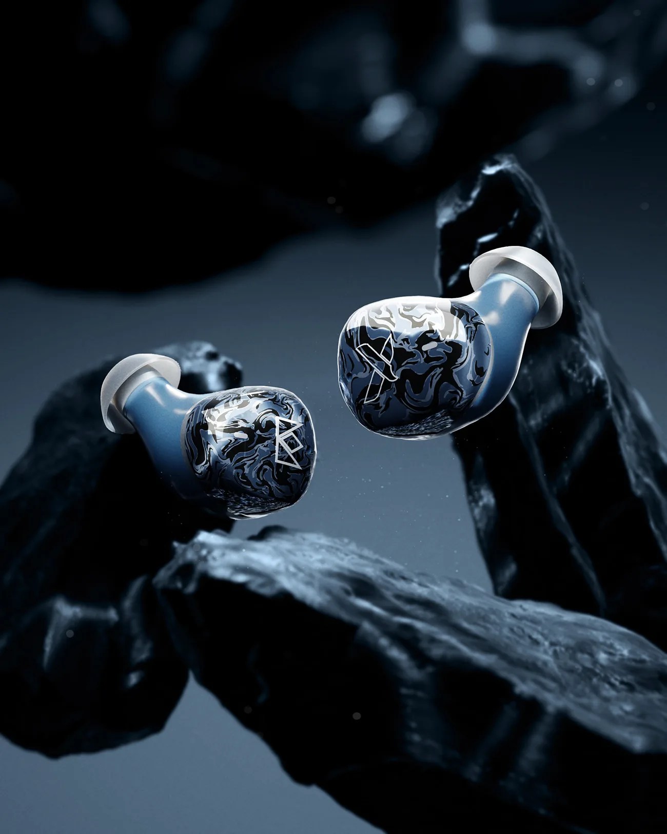 Pair of blue wireless earbuds with marbled black and white faceplates floating above dark textured rocks.
