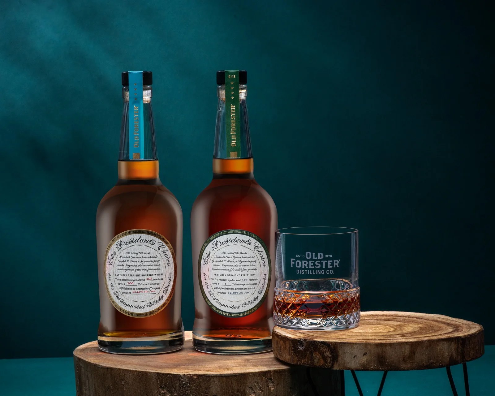 Two bottles of Old Forester whiskey and a glass with whiskey on wooden round stands against a dark teal background.