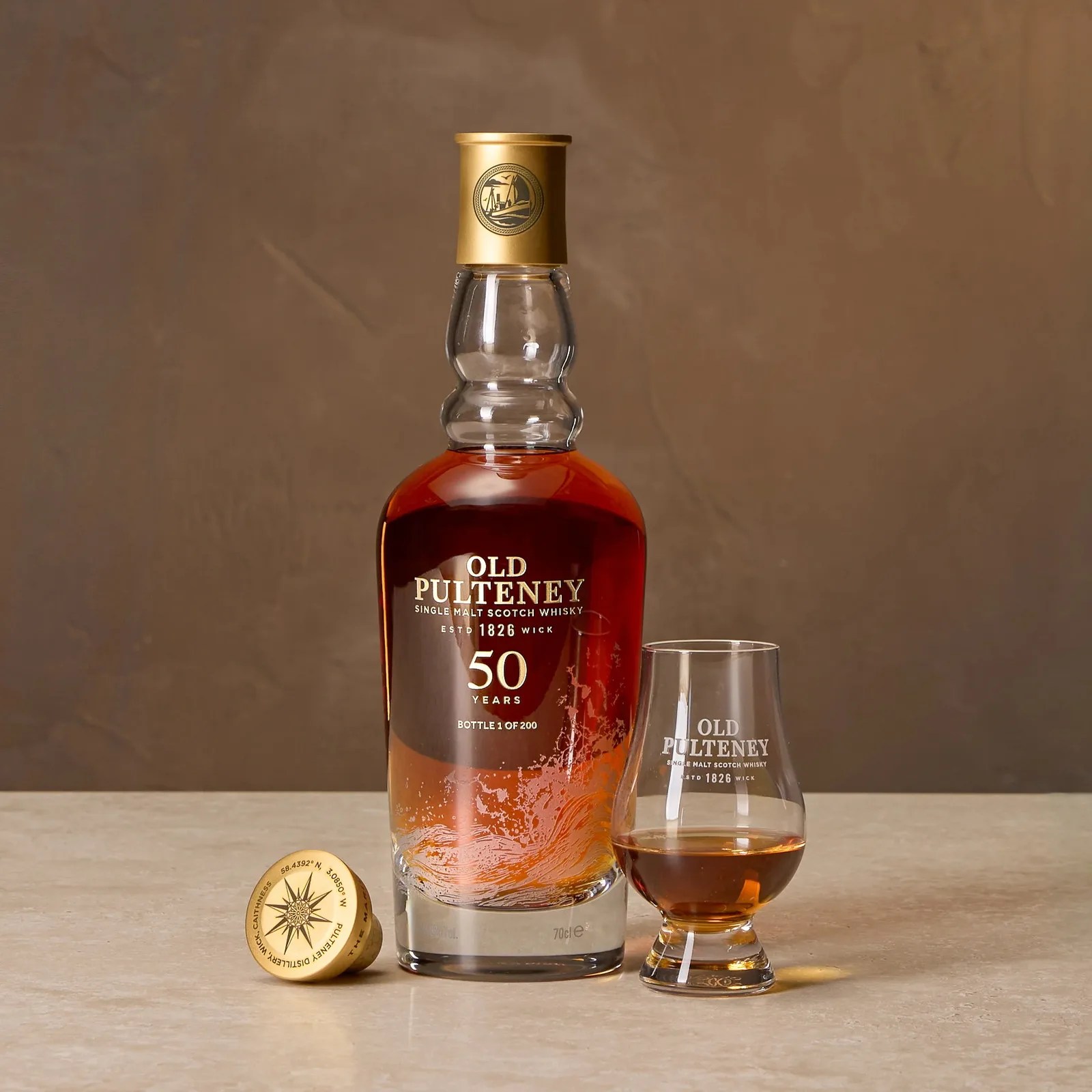 Bottle and glass of Old Pulteney 50 Years single malt Scotch whisky on a beige surface with a brown background.