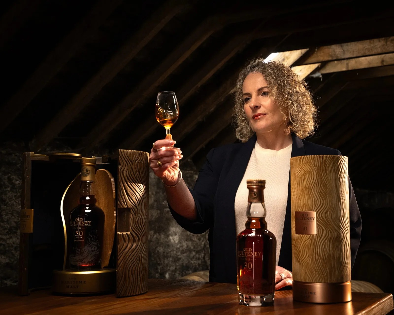 Person holding a glass of amber whiskey with Old Pulteney whiskey bottles and wooden packaging on a table.
