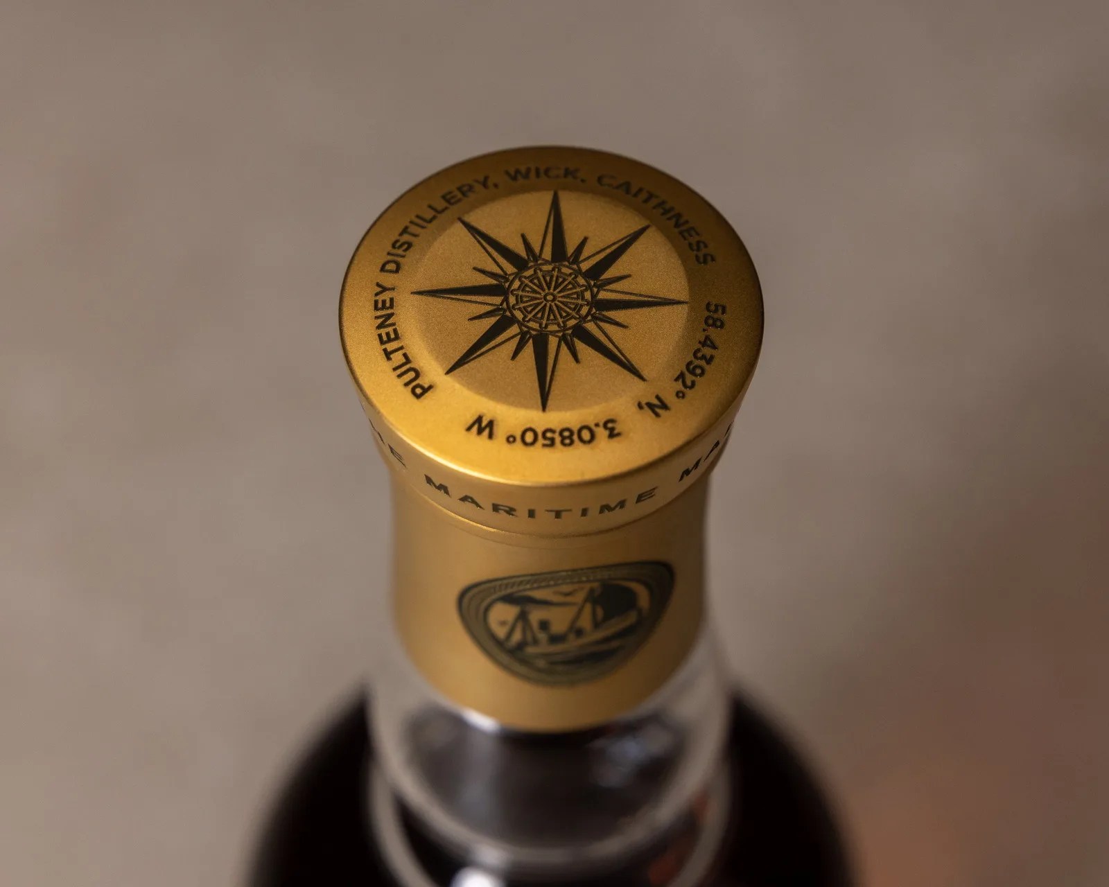 Gold bottle cap with a black compass rose design and text reading "Pulteney Distillery, Wick, Caithness" and coordinates.