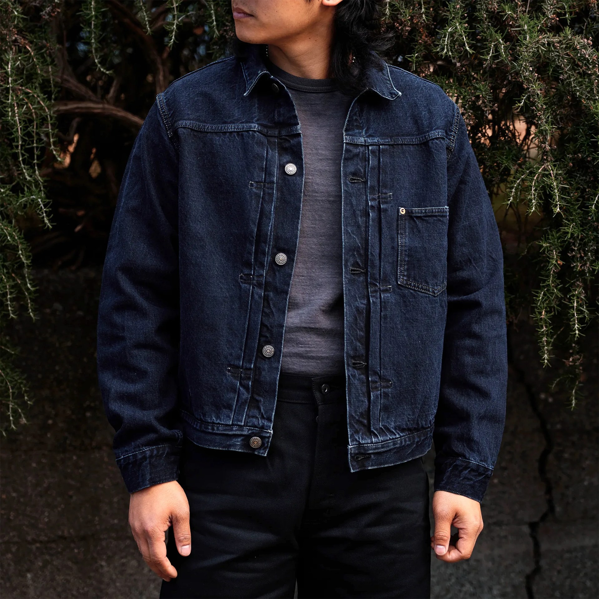 Person wearing a dark blue denim jacket over a gray shirt and black pants, standing outdoors near greenery.