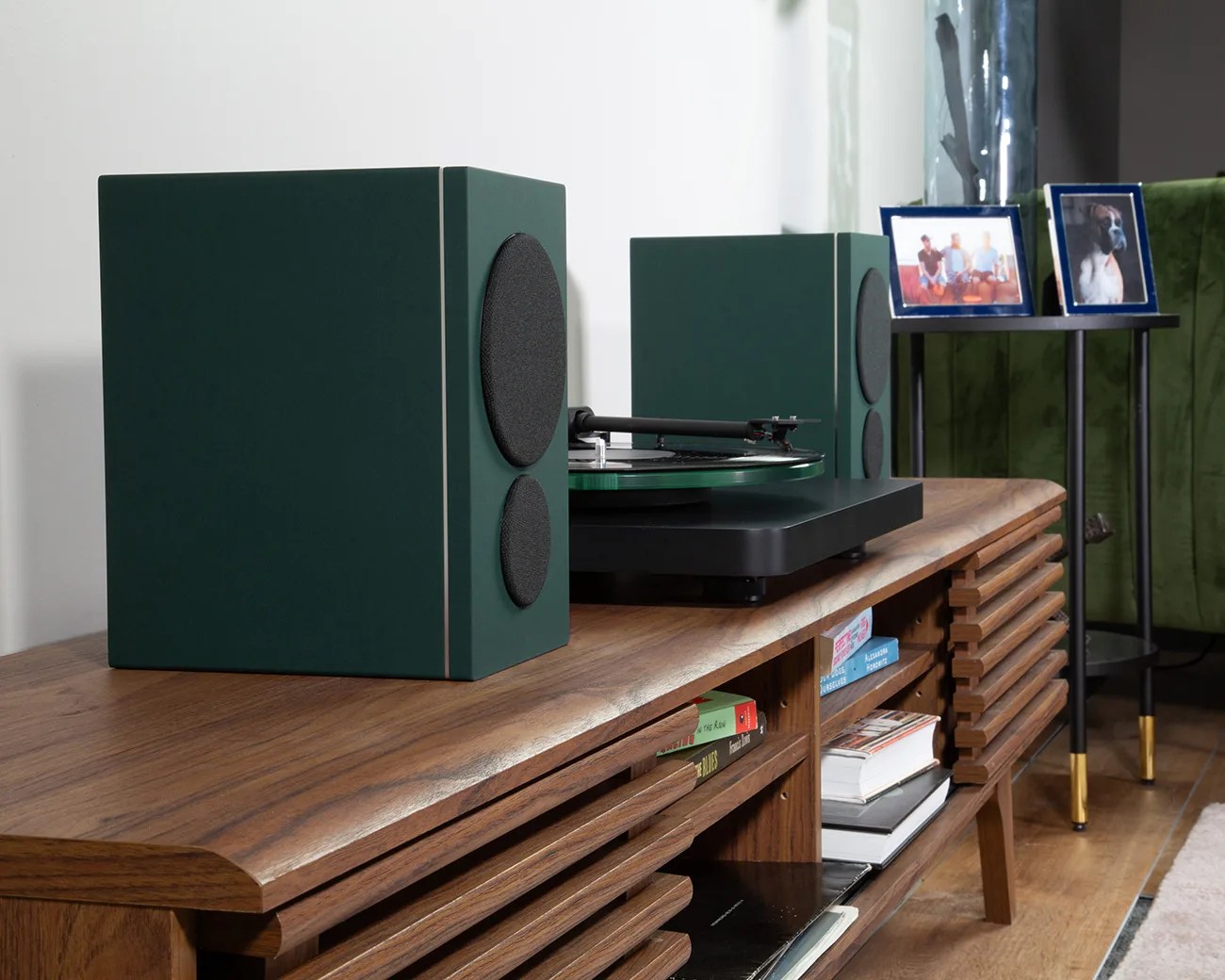 Pair of dark green bookshelf speakers with black speaker covers on a wooden media console next to a turntable.