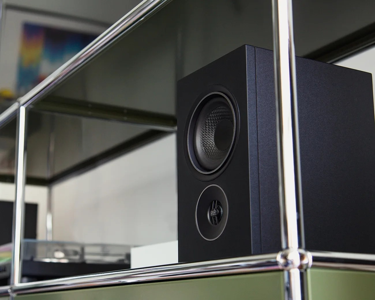 Black bookshelf speaker with textured woofer and tweeter on a glass and chrome shelf.