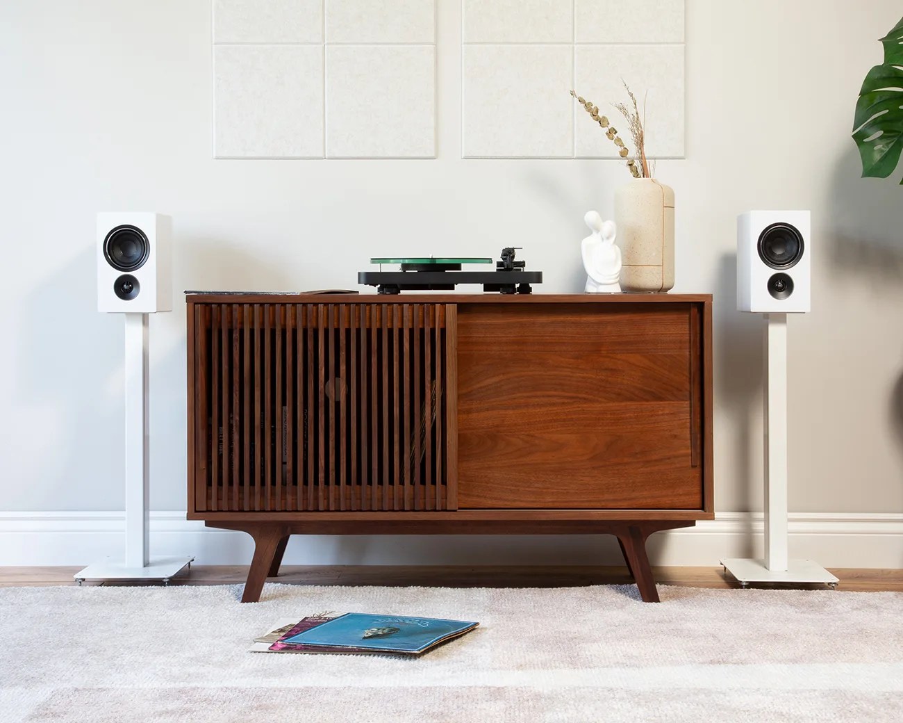 Mid-century modern wooden media console with slatted door, turntable on top, white speakers on stands, and vinyl records on carpet.