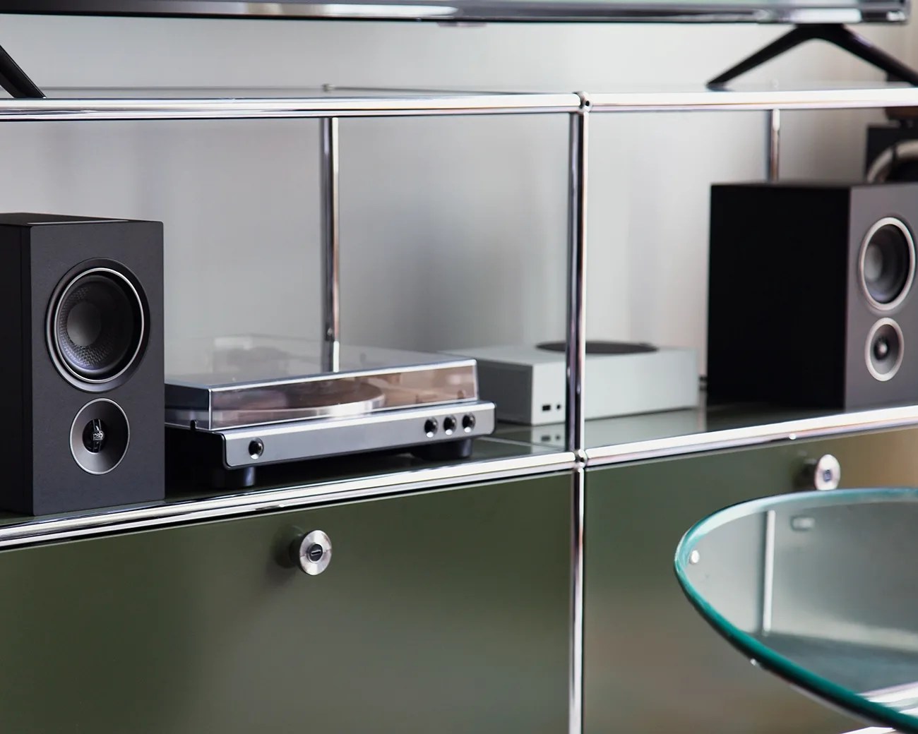 Black bookshelf speakers and a silver turntable on a glass and metal shelving unit above green cabinets.