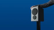 Gray rectangular speaker with two black circular drivers held by a hand against a blue background.