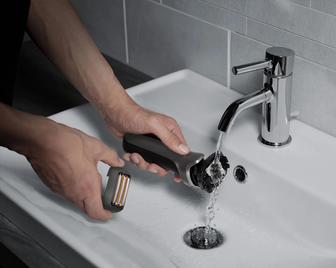 Person rinsing a black electric shaver under running water in a white sink with a chrome faucet.