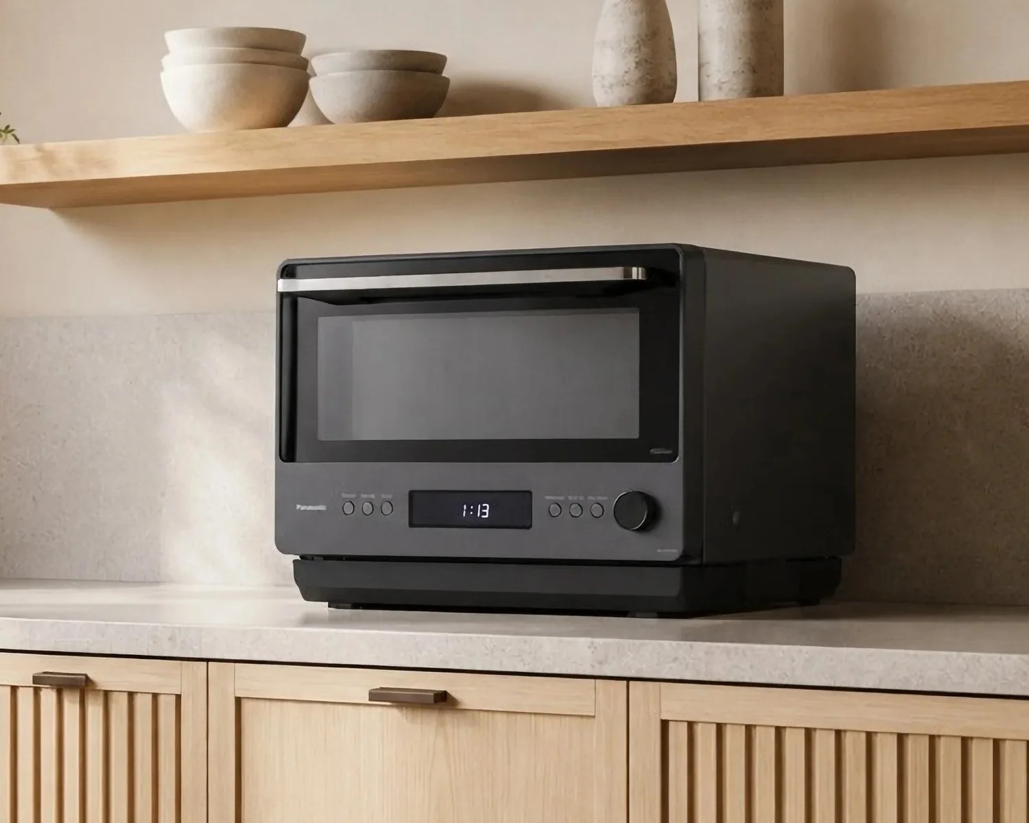 Black Panasonic microwave oven with digital display on a kitchen countertop under a wooden shelf with bowls and vases.