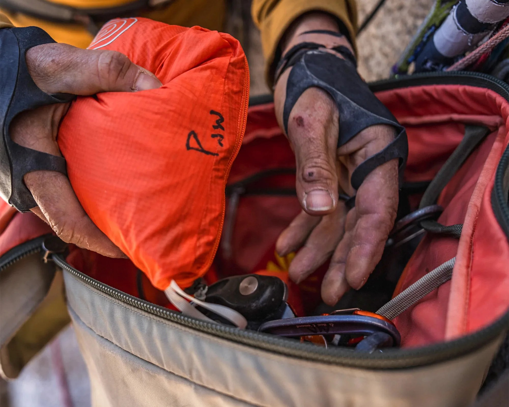 Hands wearing black fingerless gloves holding an orange stuff sack with climbing gear inside a gray and red bag.