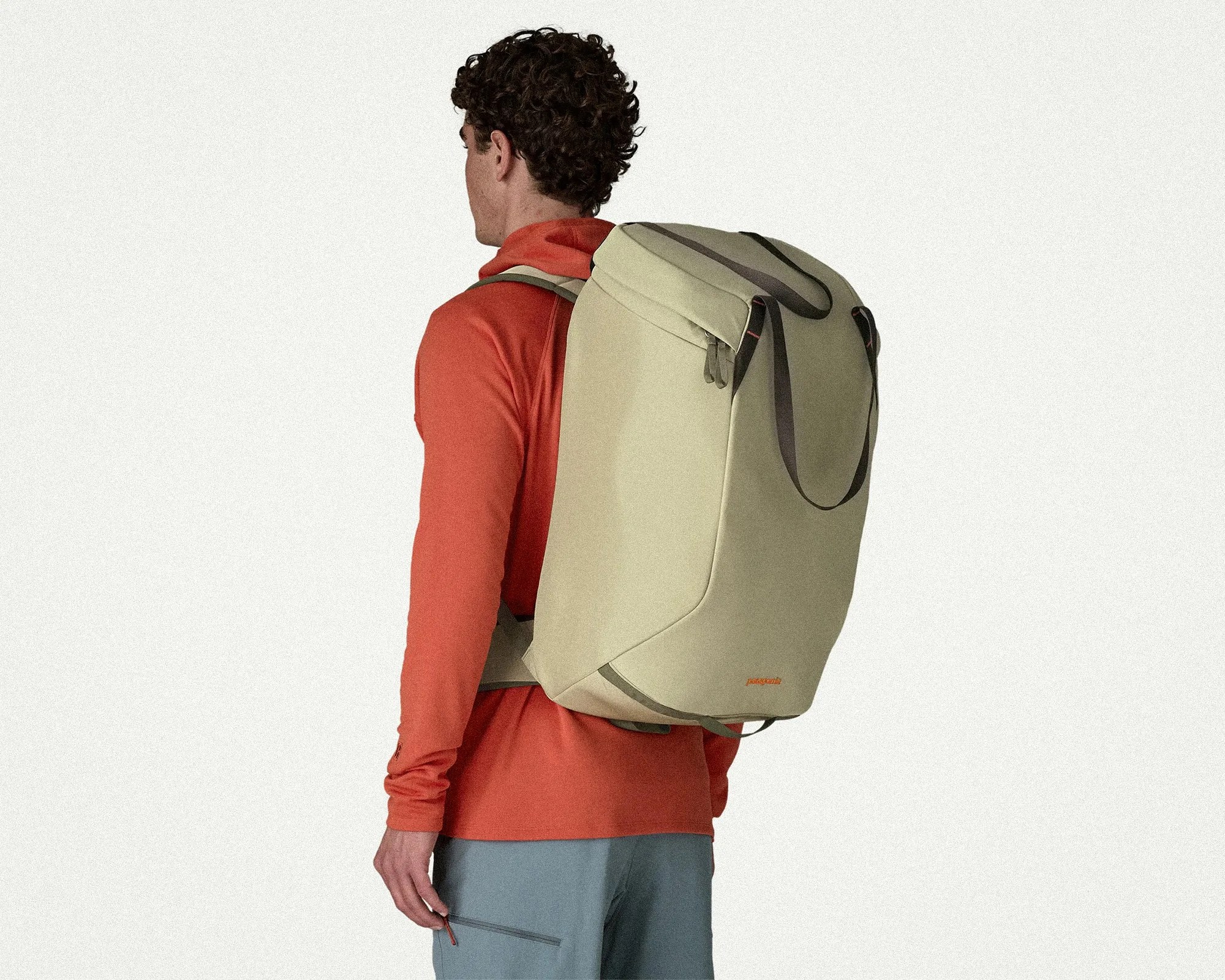 Beige rectangular backpack with black straps worn by a person in an orange long-sleeve shirt and blue pants.