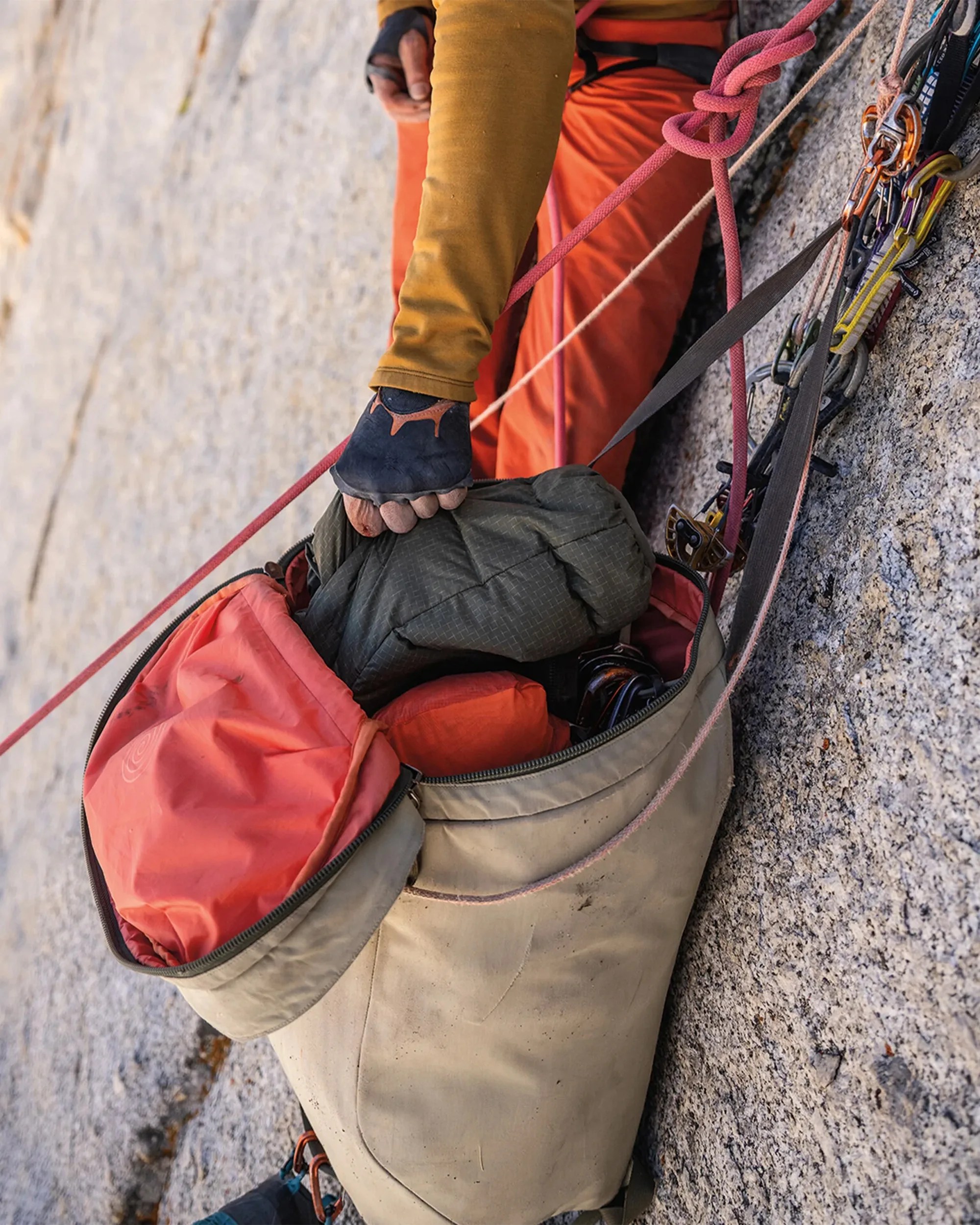 Beige and orange climbing haul bag attached to rock with climbing gear and a hand in a fingerless glove reaching inside.