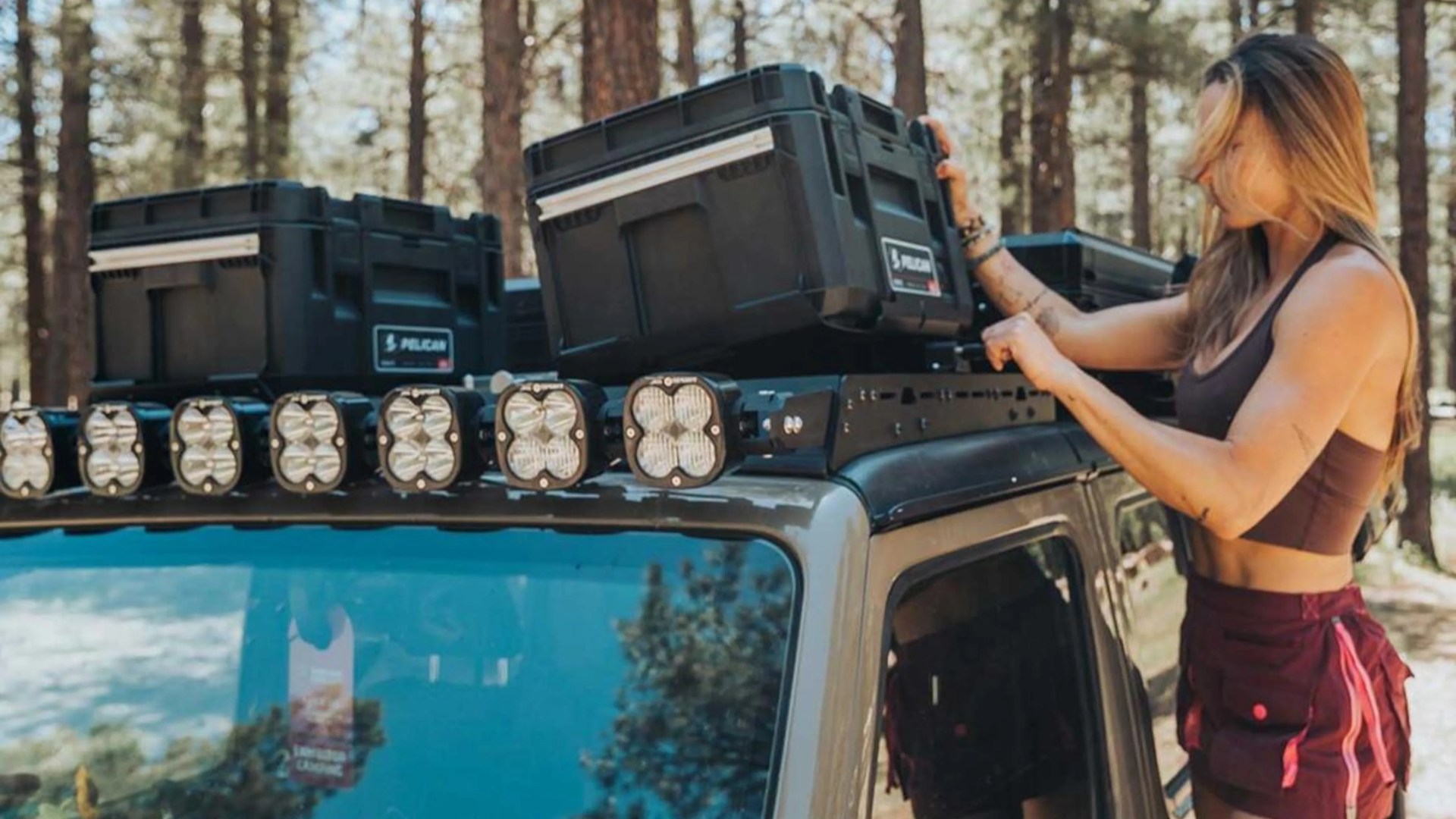 Woman securing black Pelican storage cases on a vehicle roof rack with multiple LED lights.