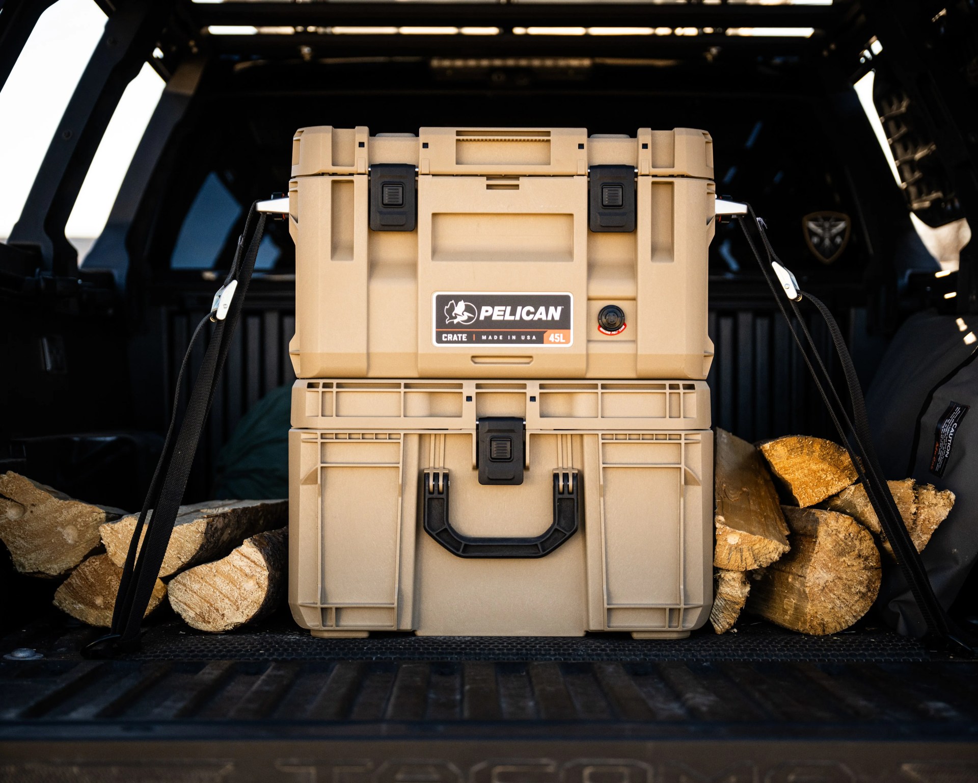 Tan Pelican 45L crate with black latches and handle, secured with straps in a vehicle trunk with firewood.