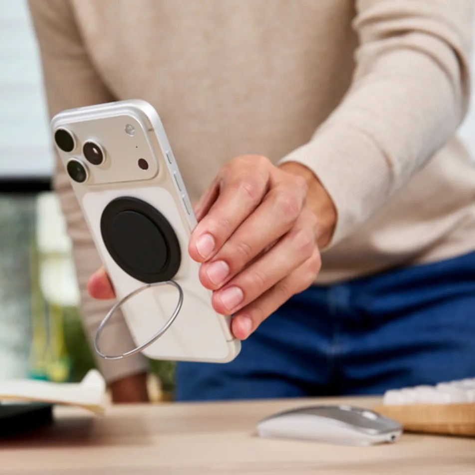 White smartphone with a black circular grip and metal ring held above a wooden desk with a wireless mouse.