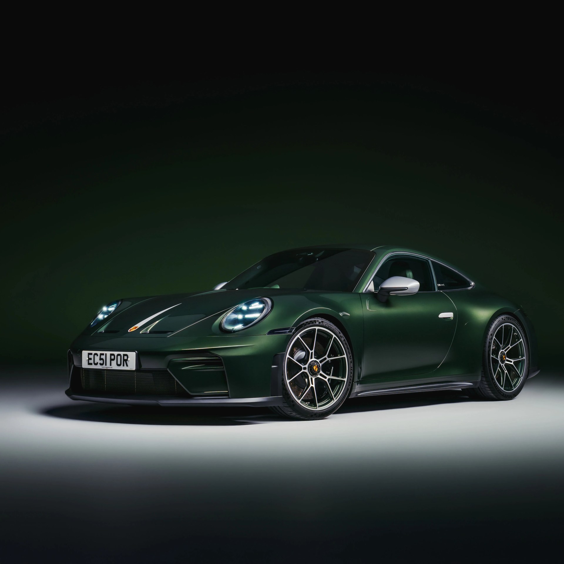 Dark green Porsche sports car with illuminated headlights and silver side mirrors on a gray surface.