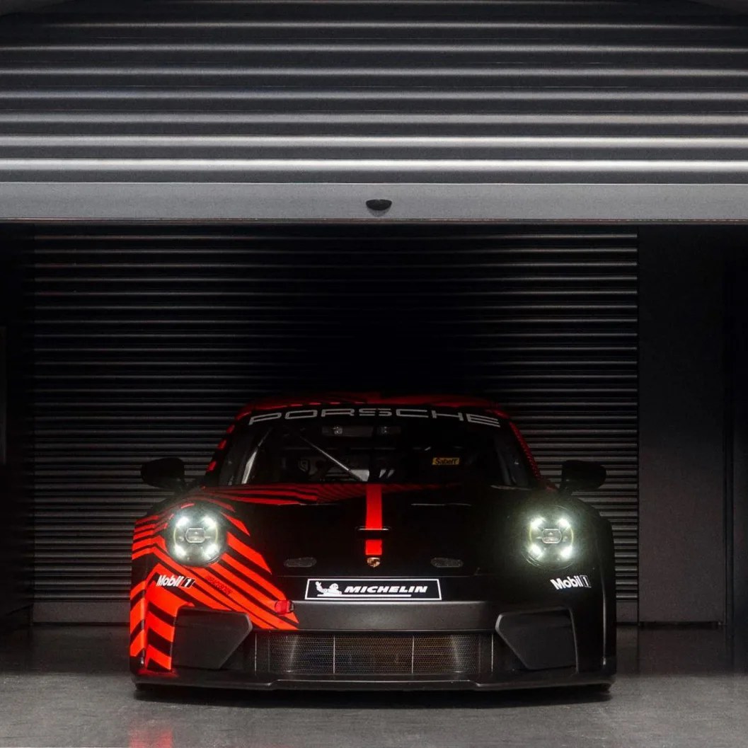 Black and red Porsche race car with headlights on parked in front of a closed metal shutter.