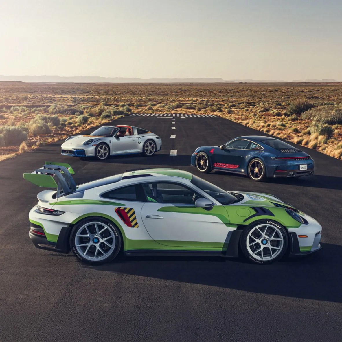 Three Porsche sports cars on a desert runway, including a white and green Porsche 911 GT3 RS in the foreground.