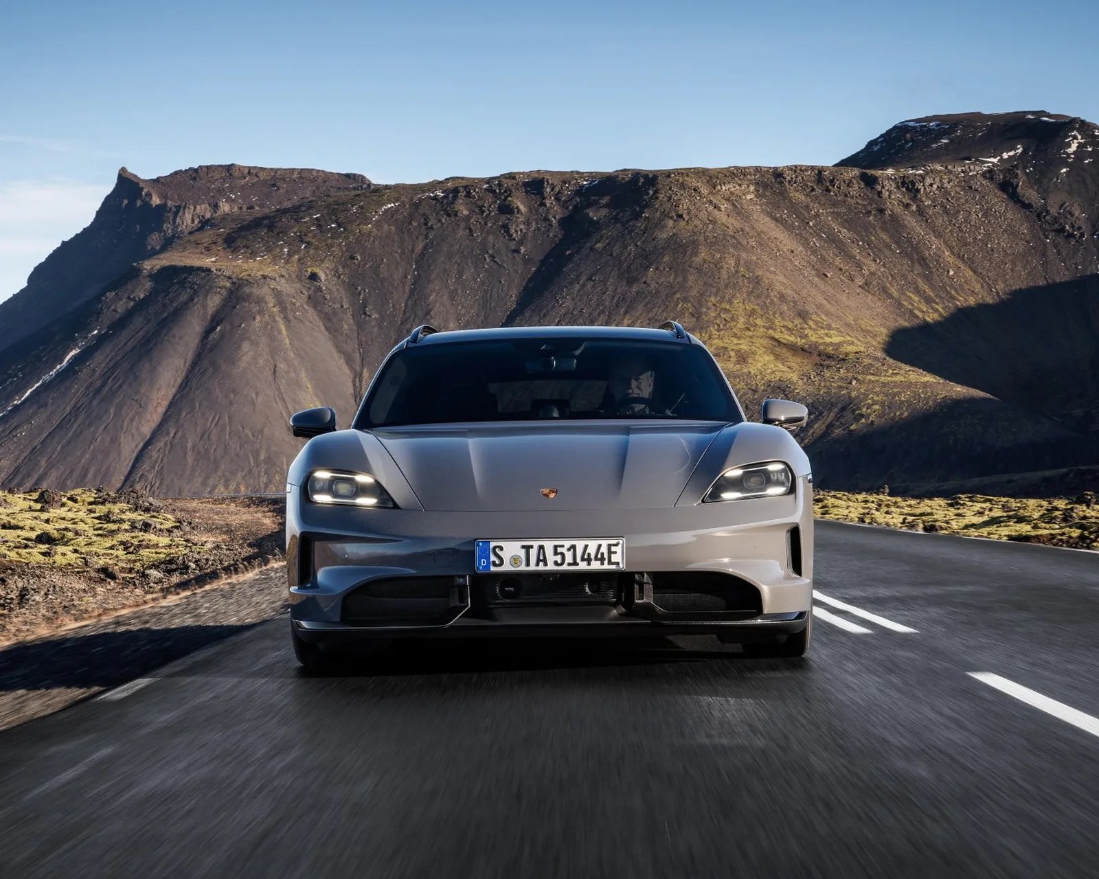Gray Porsche Taycan with headlights on driving on a mountain road with rocky hills in the background.