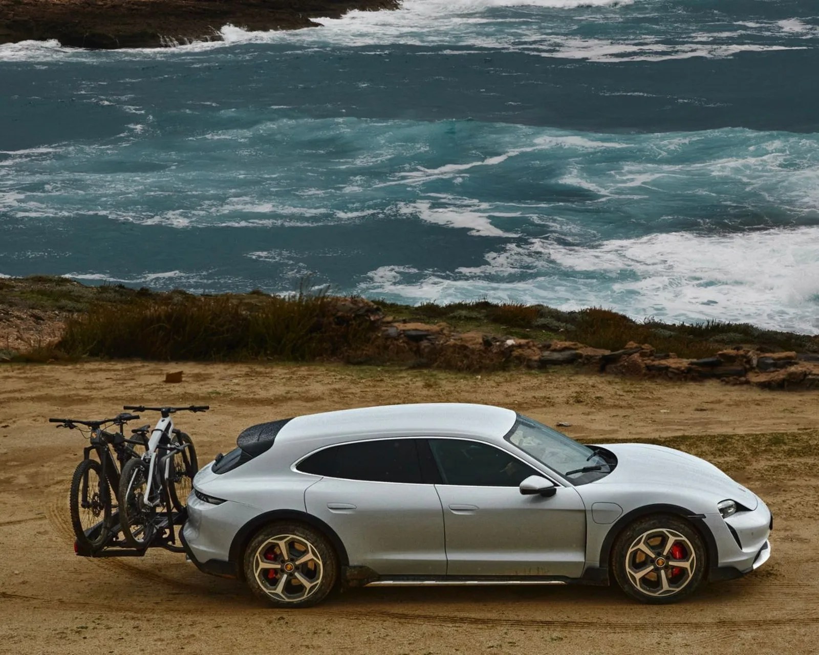 Silver electric SUV with two bicycles mounted on a rear bike rack parked on a dirt road by the ocean.