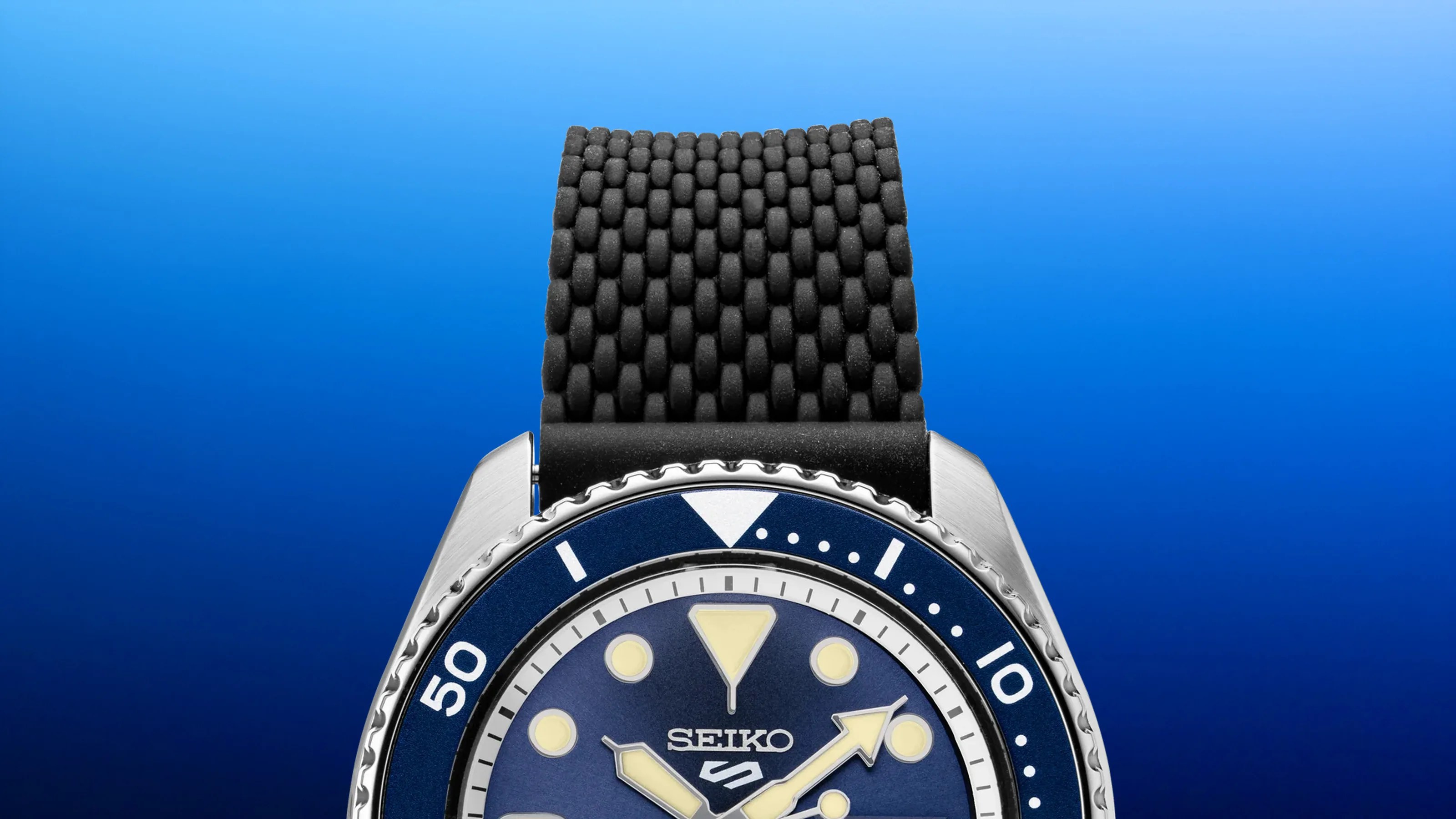 Close-up of a Seiko watch with a blue dial, luminous markers, and a textured black rubber strap.