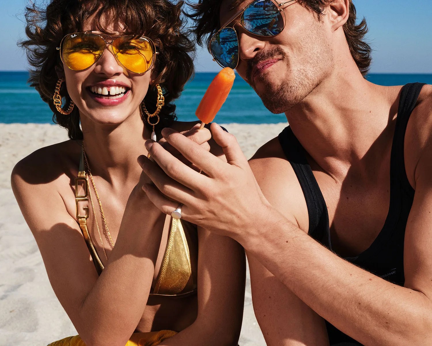 Close-up of a woman in a gold bikini and yellow sunglasses and a man in a black tank top with blue sunglasses holding an orange popsicle at the beach.