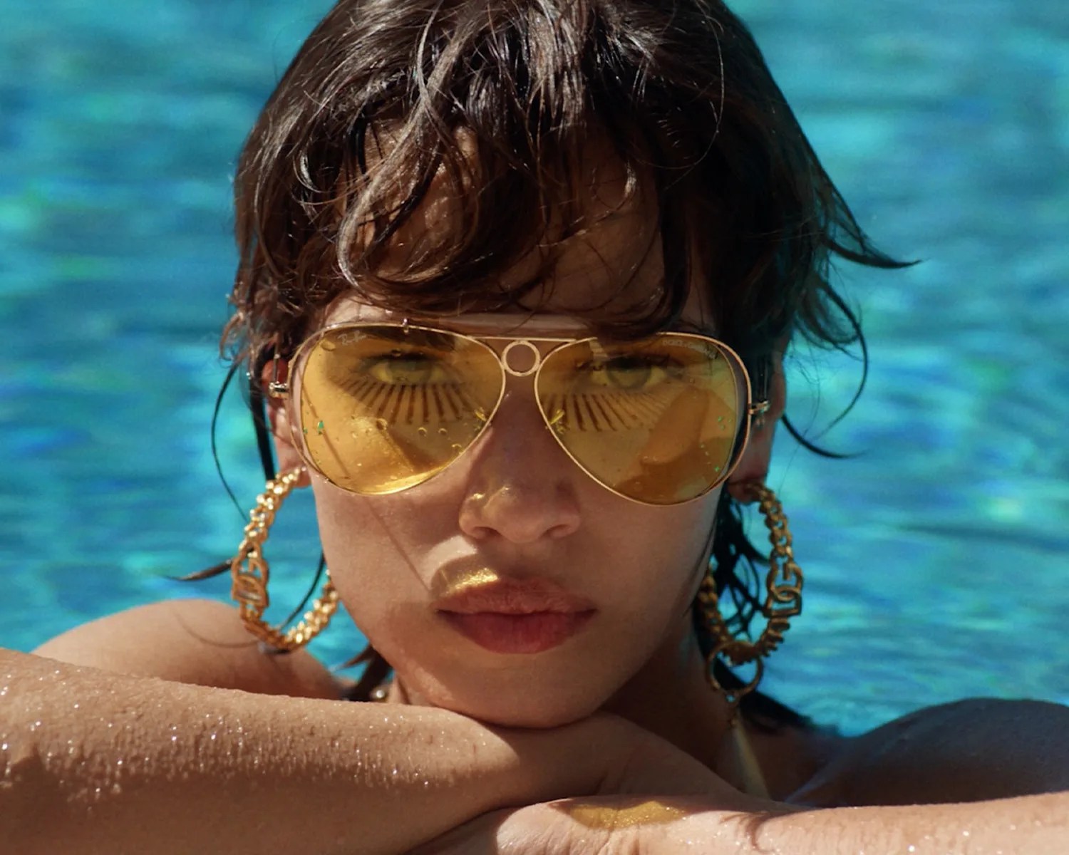 Close-up of a person wearing large gold hoop earrings and yellow-tinted aviator sunglasses in a pool.