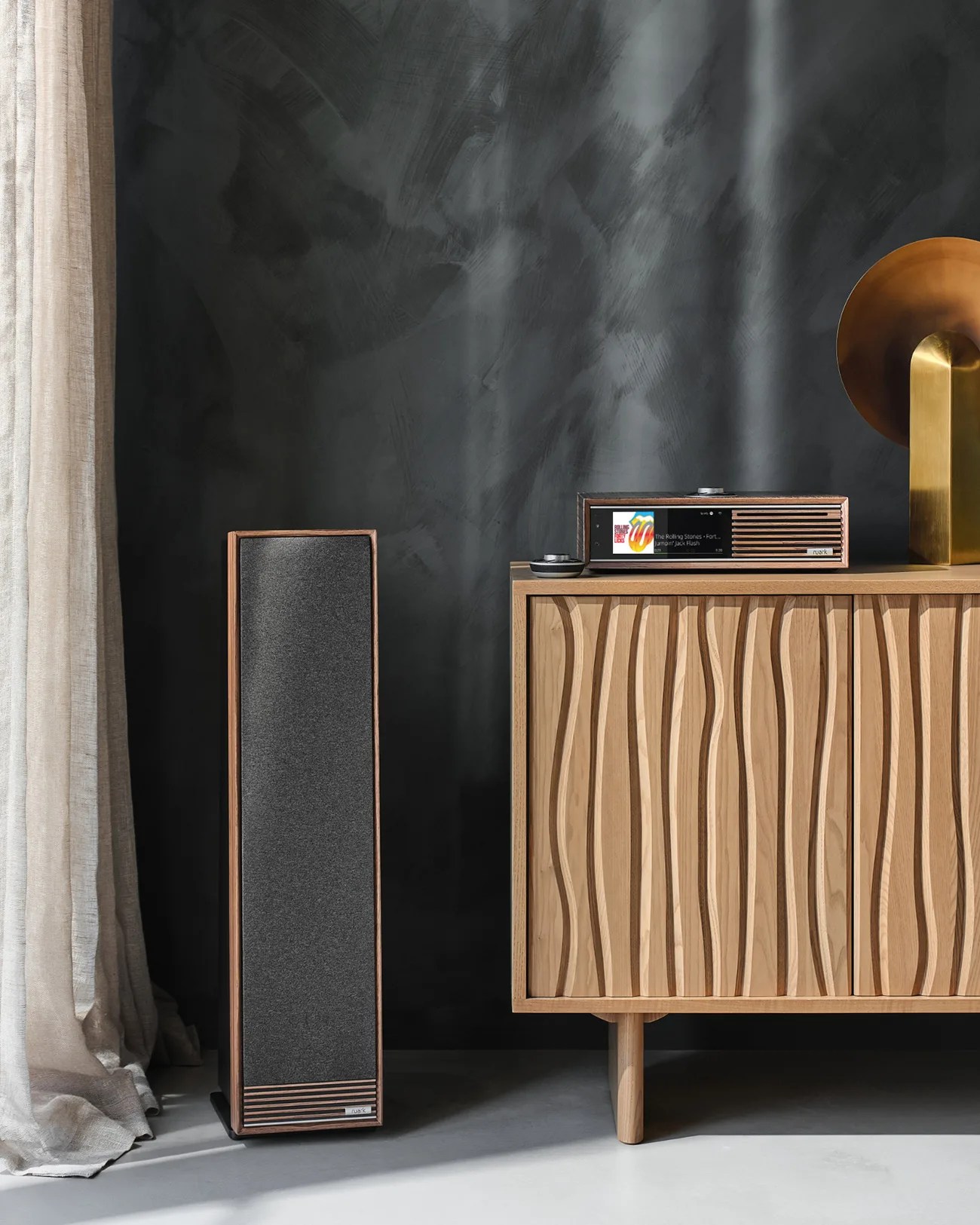 Tall wooden speaker with dark gray fabric front next to a wooden cabinet with wavy carved doors and a matching audio system on top.