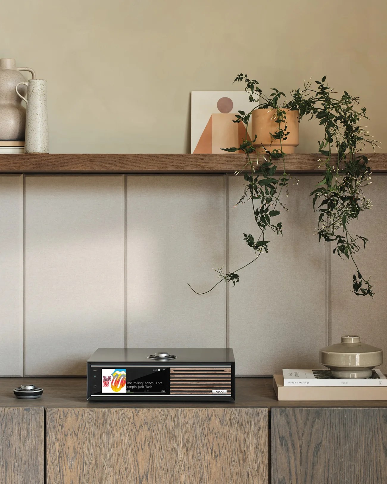 Black rectangular audio player with wooden slats and digital display on a wooden cabinet with beige wall paneling and decorative vases.