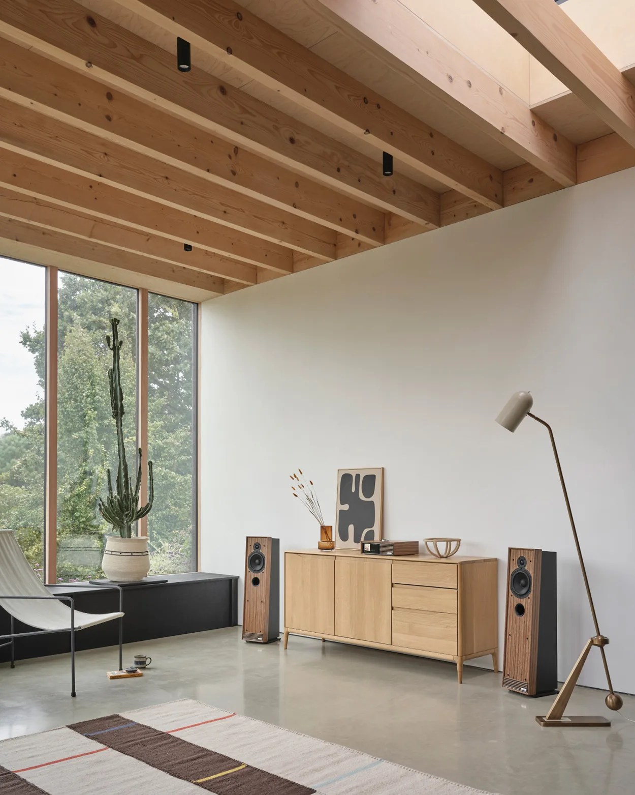 Minimalist living room with wooden ceiling beams, light wood sideboard, tall cactus in a pot, and floor lamp.