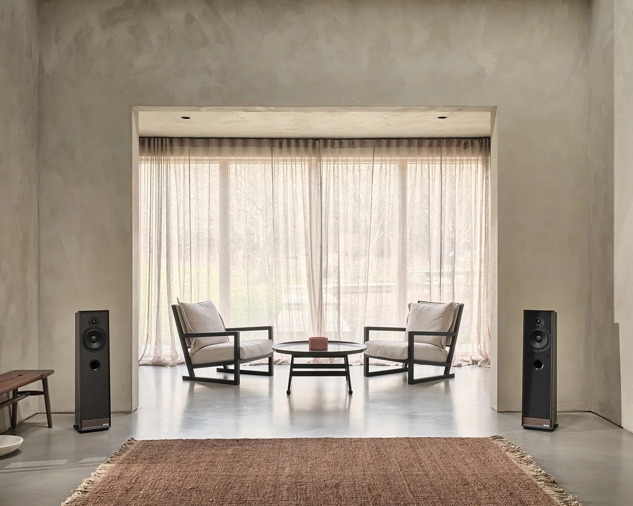 Minimalist living room with two beige cushioned armchairs, a round black coffee table, and two black floor speakers.