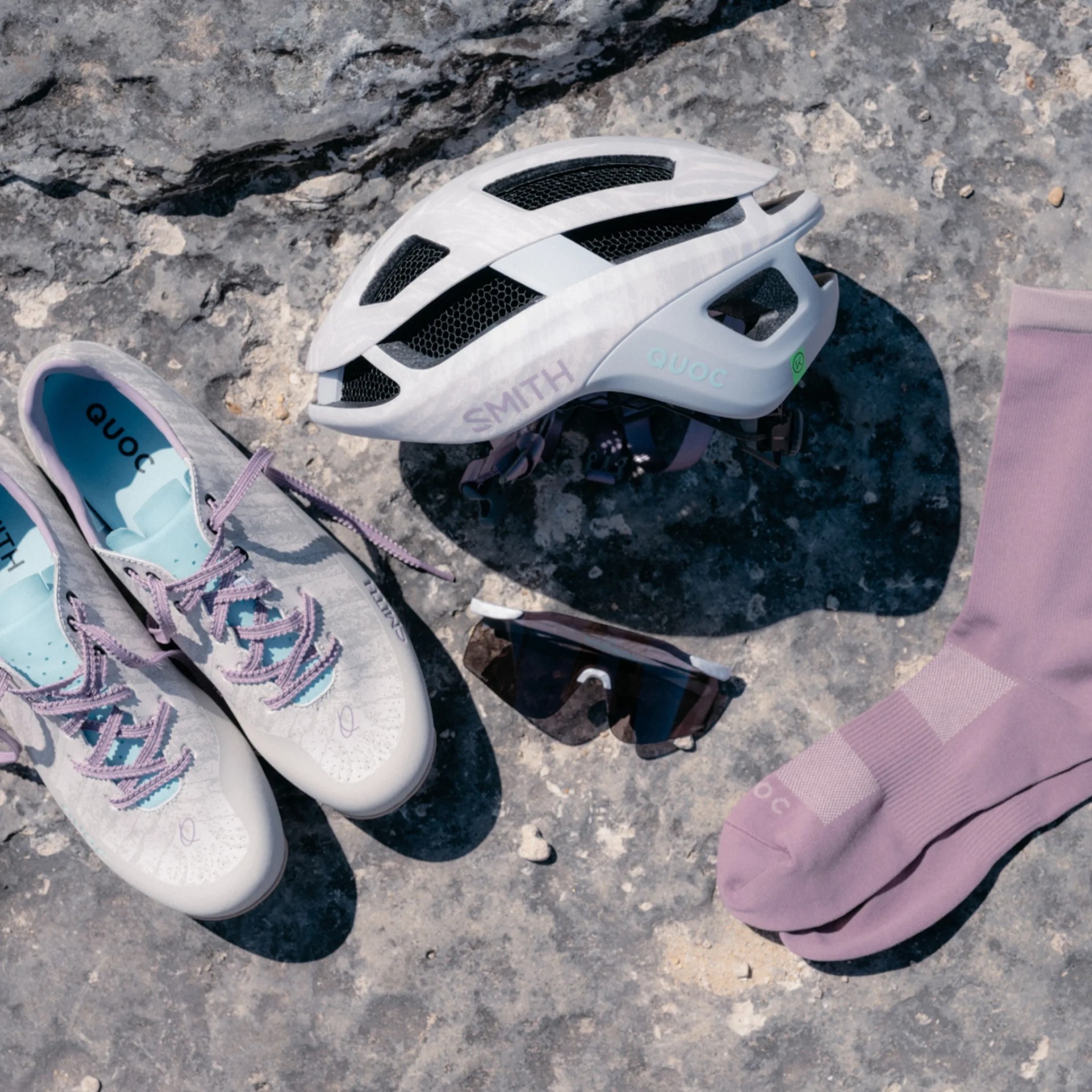 Light gray cycling shoes with purple laces, white and black helmet, black sunglasses, and purple socks on rocky ground.