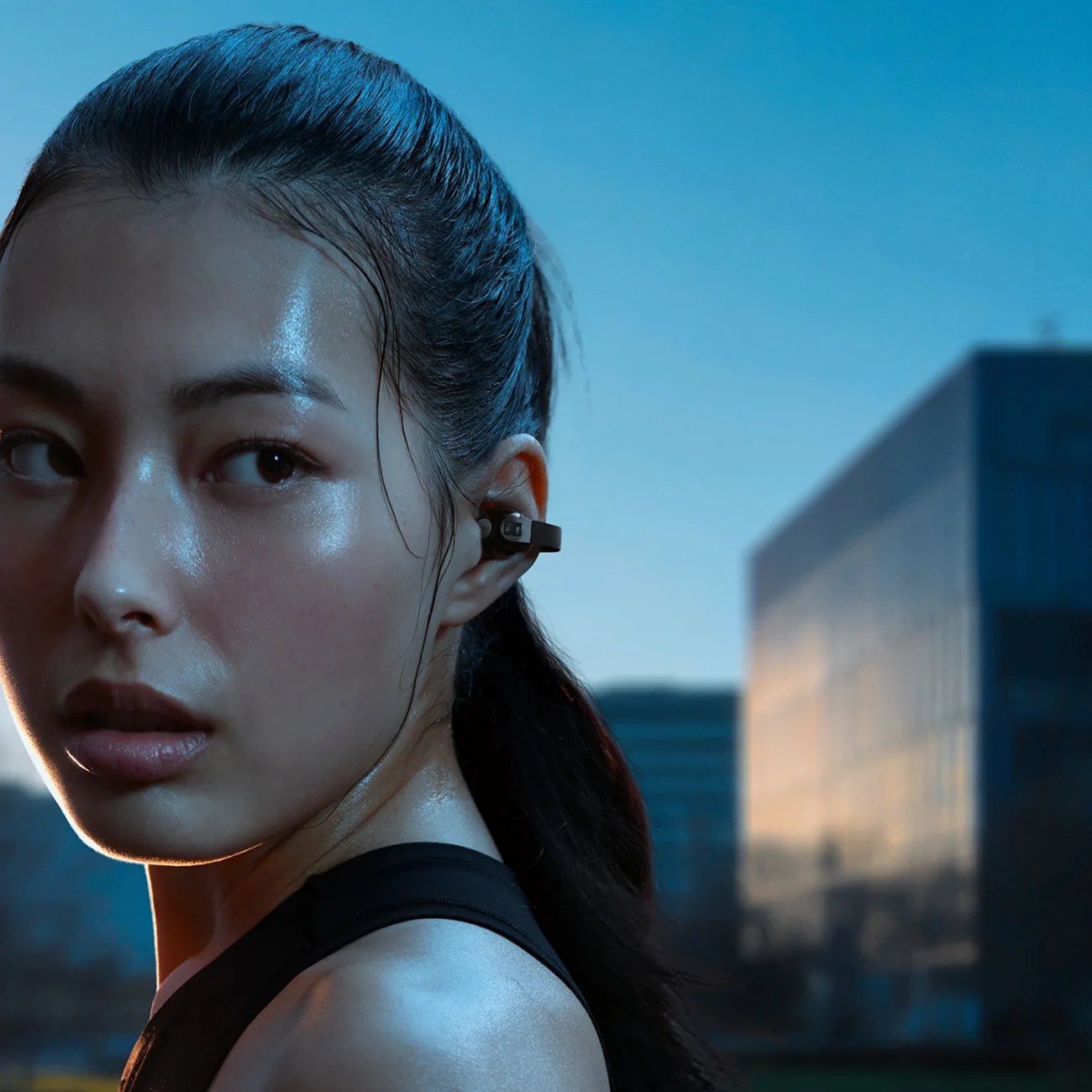 Close-up of a person wearing a black wireless earbud with a hook, outdoors at dusk.