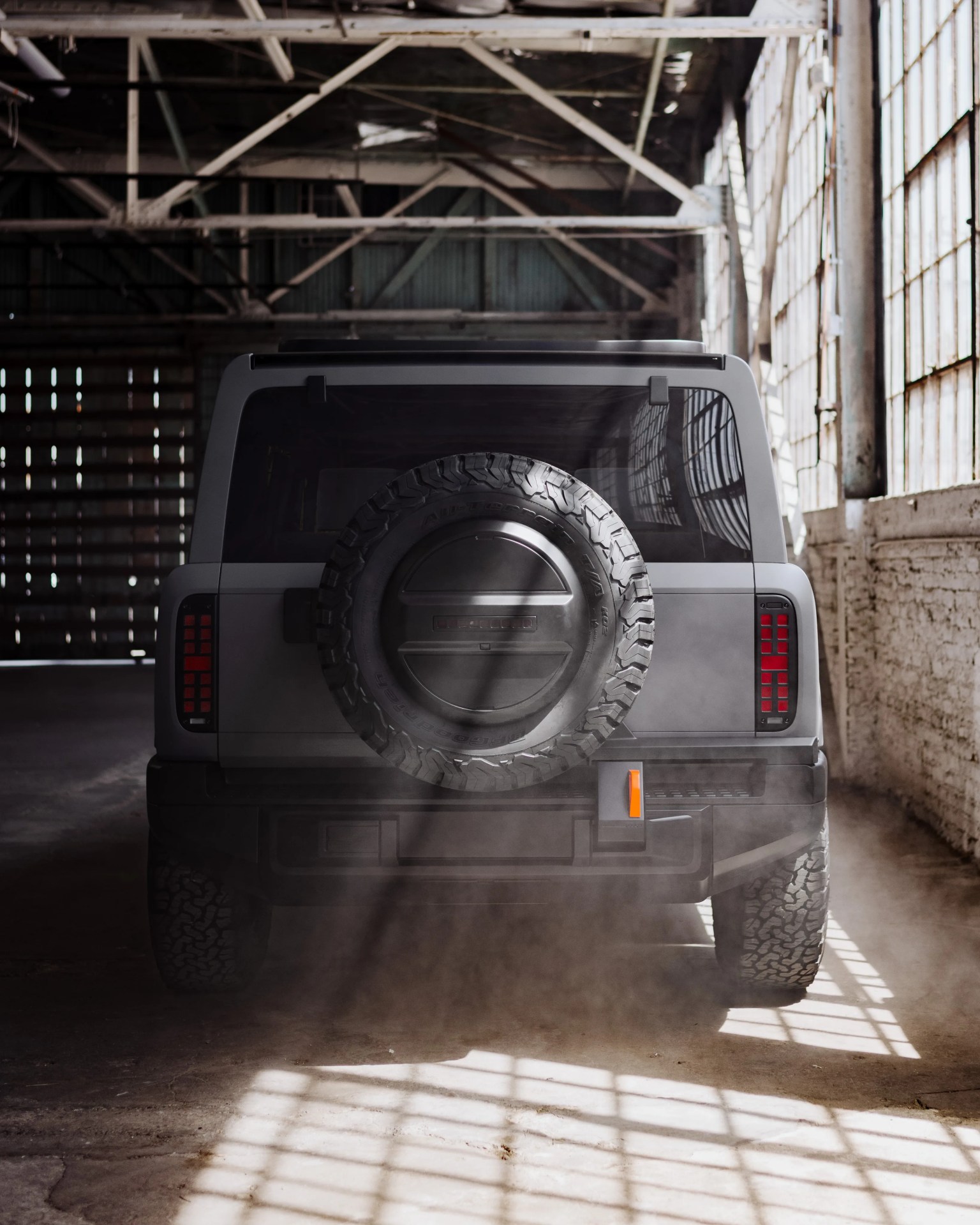 Rear view of a gray off-road vehicle with a spare tire mounted on the back, inside a sunlit industrial warehouse.