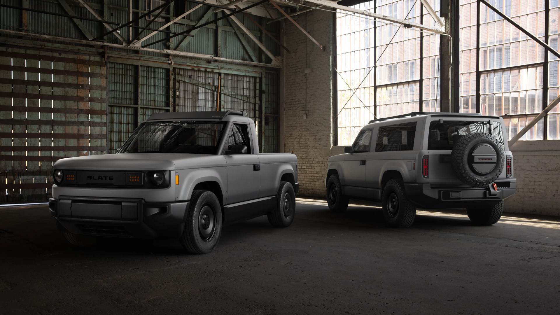 Two gray electric SUVs, one a pickup truck and the other a sport utility vehicle, parked inside a warehouse with large windows.