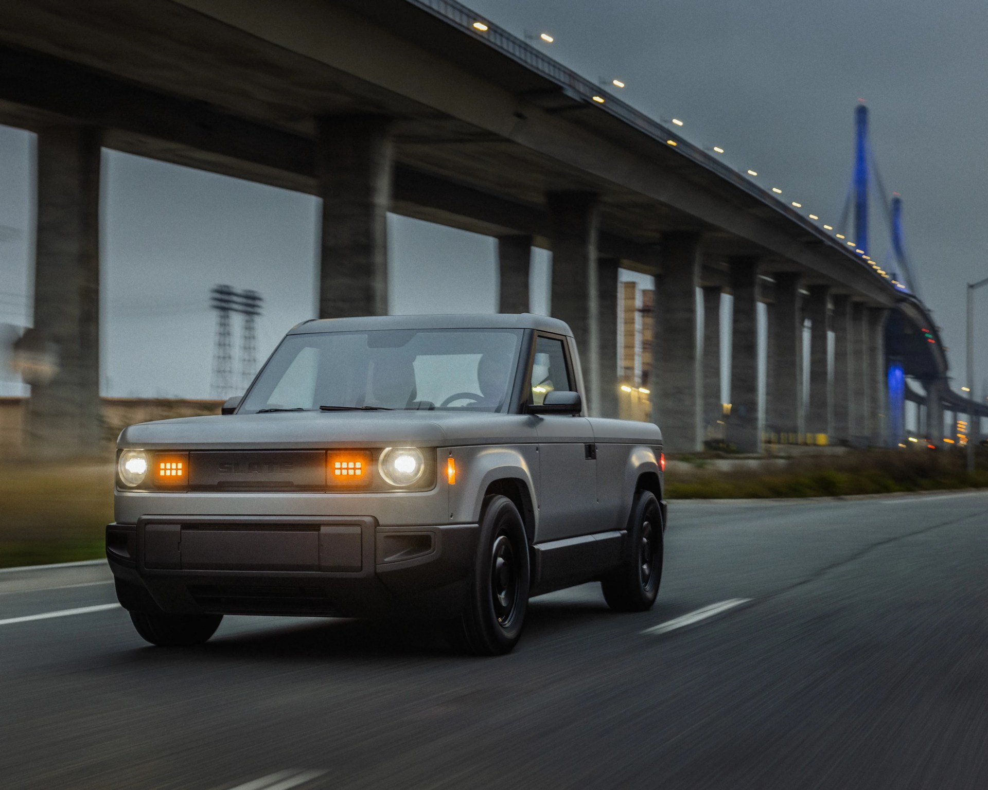 Gray electric pickup truck driving on a road under a large concrete bridge at dusk.