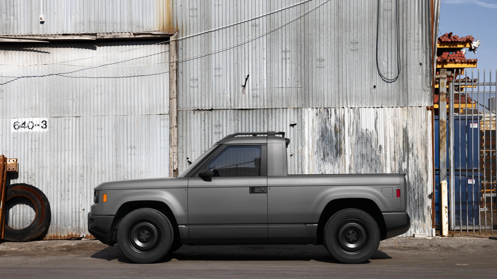 Matte gray compact pickup truck parked in front of a corrugated metal wall with rust and peeling paint.