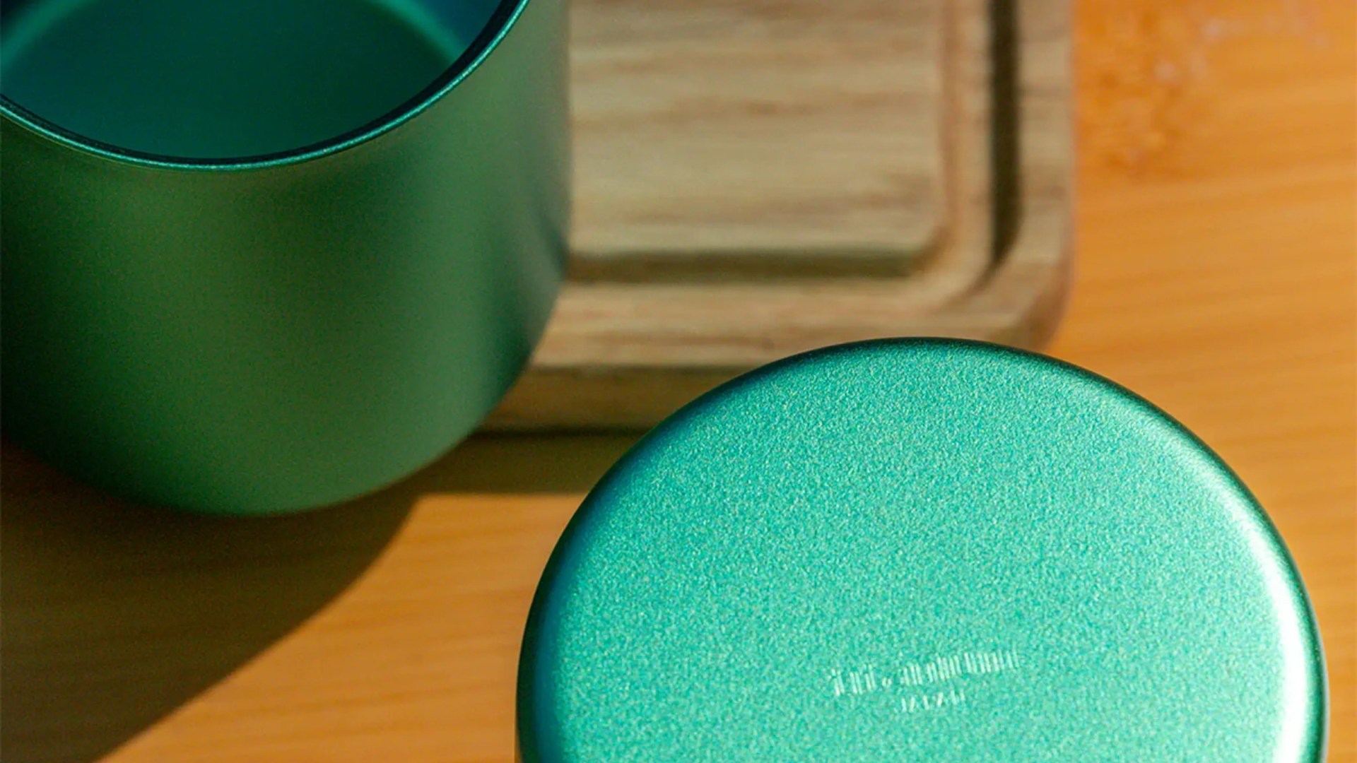 Teal metallic cylindrical container with a matching lid on a wooden surface next to a wooden tray.