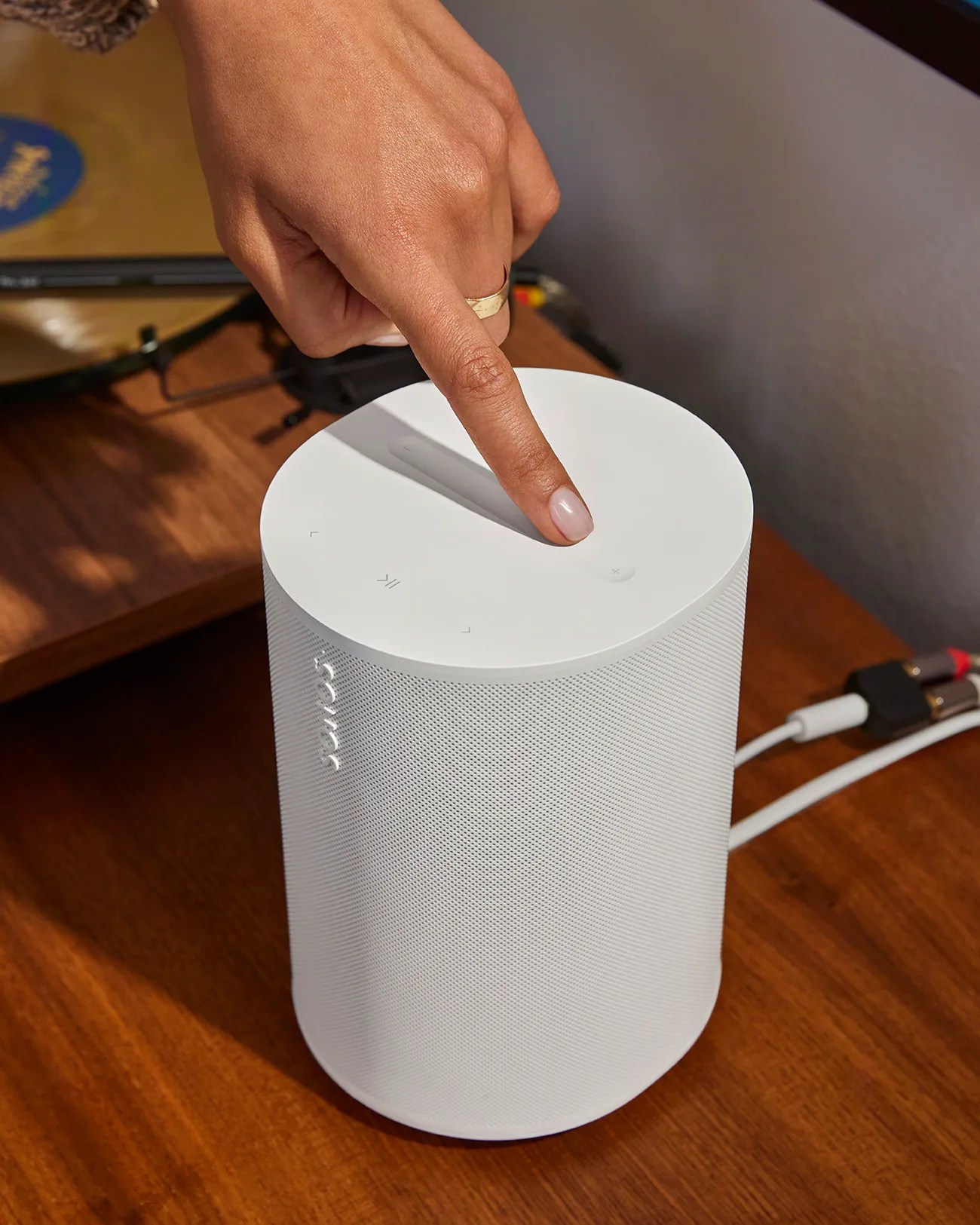 White cylindrical Sonos speaker on a wooden surface with a hand pressing the volume up button on top.