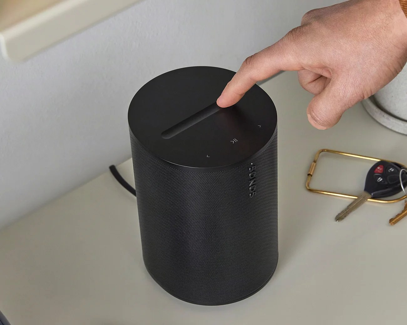 Black cylindrical Sonos speaker with touch controls on top, a hand about to press a button, on a light-colored surface.