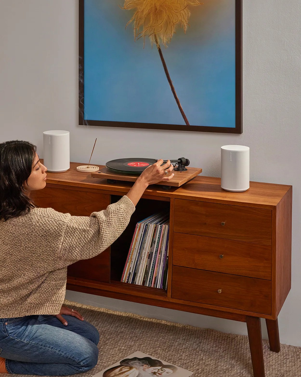 Woman adjusting a wooden turntable on a mid-century modern wooden cabinet with white Sonos speakers and vinyl records.
