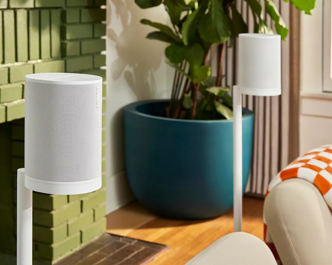 Two white Sonos speakers mounted on white stands in a living room with a green brick wall and a large blue plant pot.
