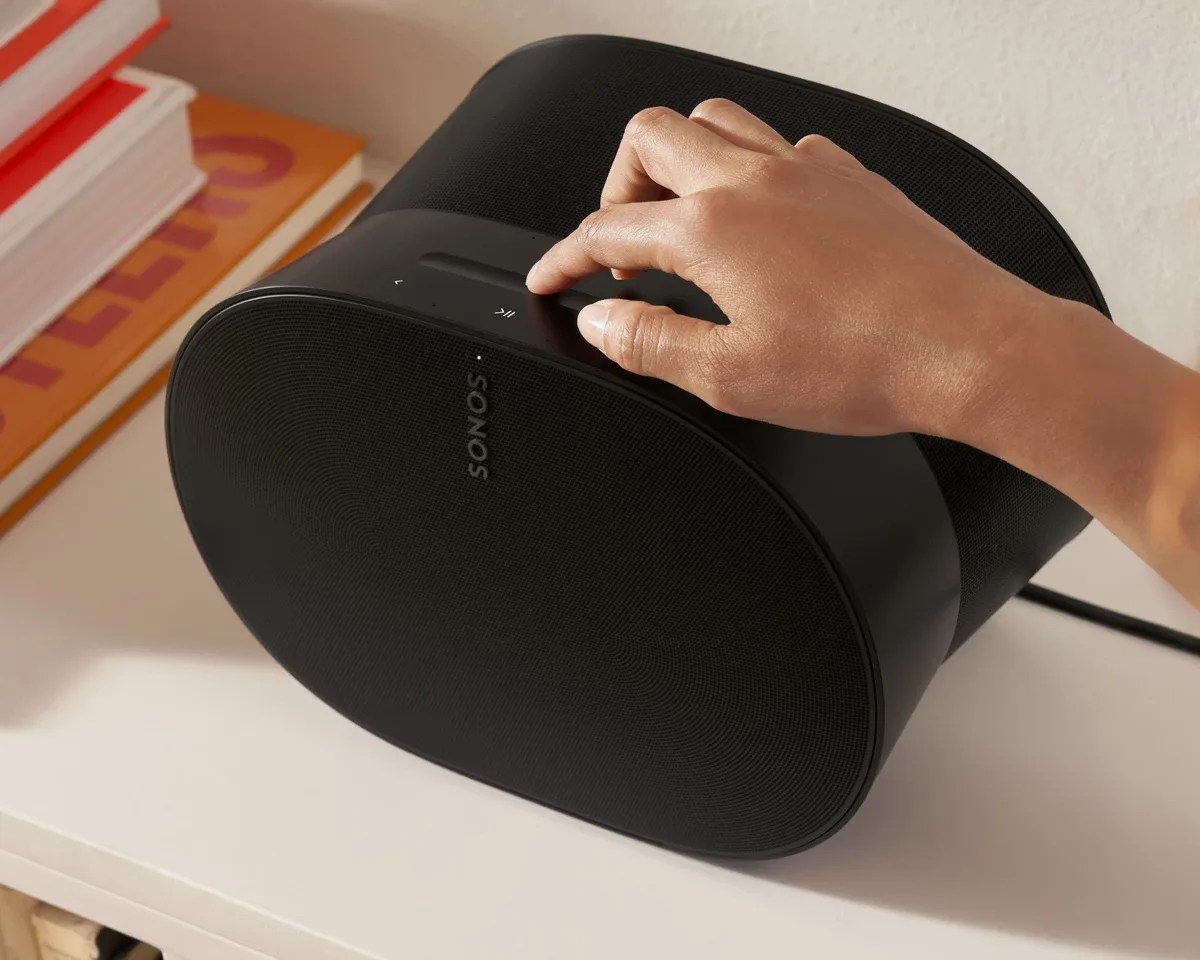 Black Sonos speaker with touch controls being operated by a hand on a white surface near stacked books.