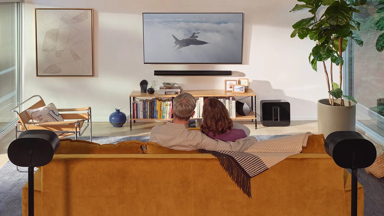 Two people sitting on a mustard yellow sofa watching a fighter jet on a wall-mounted TV in a modern living room.