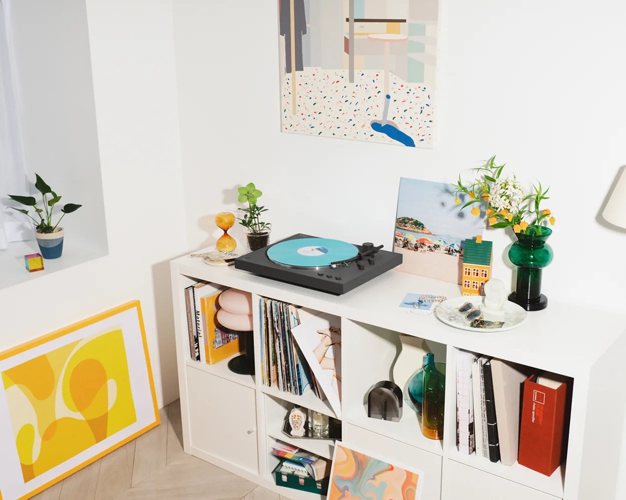 White cubby shelf with records, books, a black turntable with a blue vinyl, plants, and decorative items in a bright room.