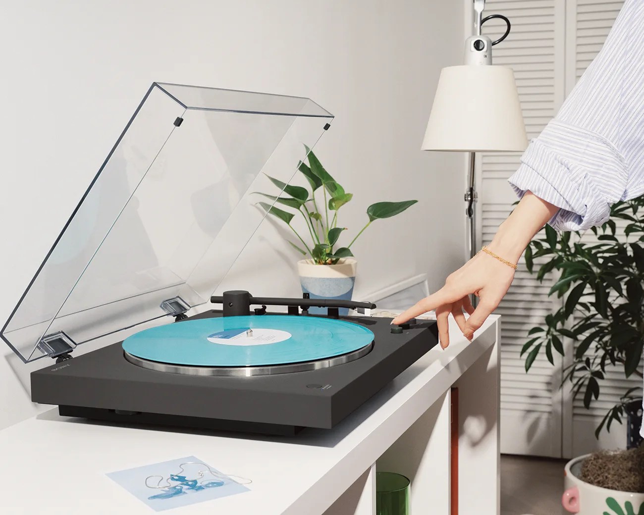 Black turntable with a blue vinyl record and a transparent dust cover on a white table, with a hand pressing a button.