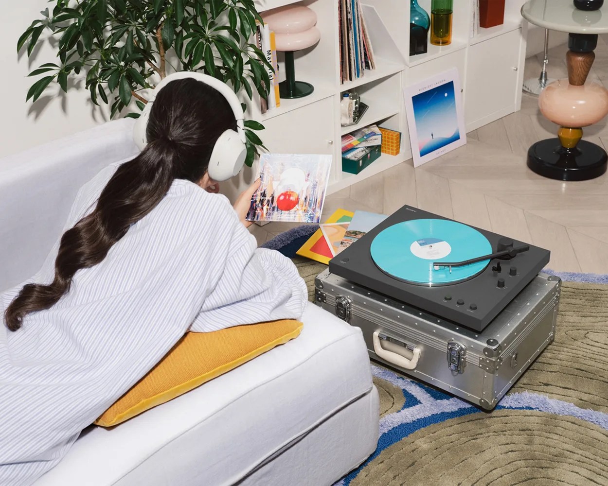 Person wearing white headphones lying on a white couch with a yellow pillow, holding a vinyl record near a black turntable with a blue record.